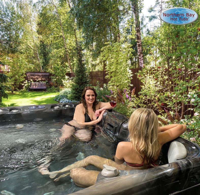 Two women relaxing in a hot tub surrounded by trees in a residential backyard